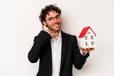 Beyaz arka planda izole edilmiş oyuncak evi tutan Kafkasyalı genç iş adamı parmaklarıyla bir cep telefonu hareketi gösteriyor..