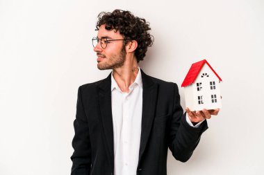 Beyaz arka planda izole edilmiş oyuncak evi tutan Kafkasyalı genç bir iş adamı gülümsüyor, neşeli ve keyifli görünüyor..