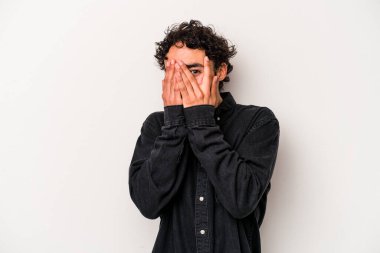 Beyaz arka planda izole edilmiş genç beyaz bir adam parmaklarının arasından korkmuş ve gergin bir şekilde göz kırpıyor..