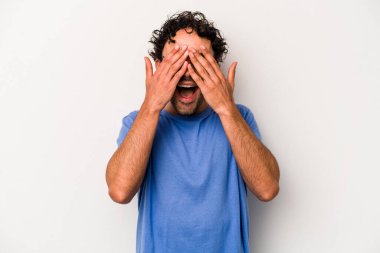 Beyaz arka planda soyutlanmış beyaz tenli genç bir adam elleriyle gözlerini kapatıyor, geniş bir gülümsemeyle bir sürpriz bekliyor..