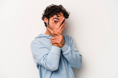 Beyaz arka planda izole edilmiş genç beyaz bir adam parmaklarının arasından korkmuş ve gergin bir şekilde göz kırpıyor..