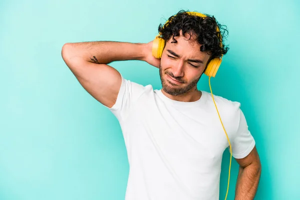 Young caucasian man listening to music isolated on blue background touching back of head, thinking and making a choice.