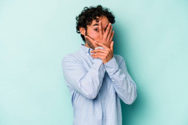 Beyaz arka planda izole edilmiş genç beyaz bir adam parmaklarının arasından korkmuş ve gergin bir şekilde göz kırpıyor..