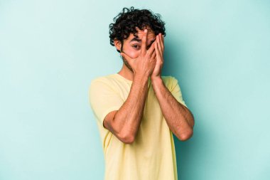 Beyaz arka planda izole edilmiş genç beyaz bir adam parmaklarının arasından korkmuş ve gergin bir şekilde göz kırpıyor..