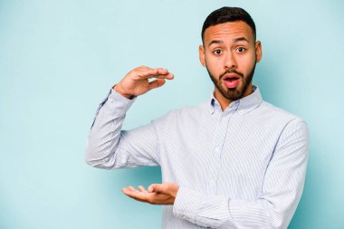 Mavi arka planda izole edilmiş genç İspanyol adam elleri arasında bir kopya alanı tutarken şok oldu ve hayrete düştü..