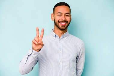 Mavi arka planda izole edilmiş genç İspanyol adam zafer işareti gösteriyor ve gülümsüyor..