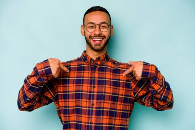 Mavi arka planda izole edilmiş genç İspanyol adam parmağını göstererek şaşırdı, geniş bir gülümsemeyle..