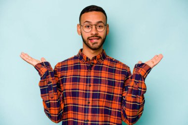 Genç İspanyol bir adam mavi arka planda izole edilmiş. Kafası karışmış ve omuz silkiyor. Kopya alanını tutmak için..