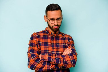 Latin asıllı genç adam mavi arka planda izole edilmiş. Mutsuz bir şekilde kameraya bakıyor. İğneleyici bir ifadeyle..