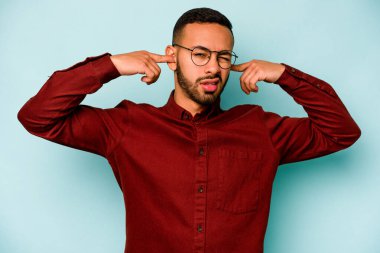 Genç İspanyol bir adam mavi arka planda izole edilmiş parmaklarıyla kulaklarını kapatıyor, gürültülü bir ortam yüzünden stresli ve çaresiz..