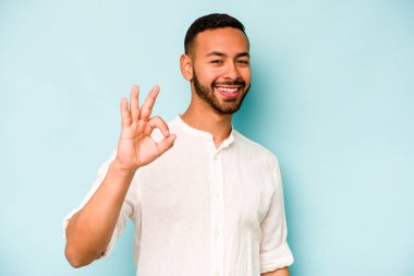 Genç İspanyol adam mavi arka planda izole edilmiş neşeli ve kendine güvenen iyi bir hareket sergiliyor..