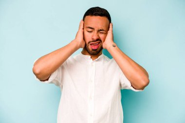 Mavi arka planda izole edilmiş genç İspanyol adam kulaklarını elleriyle kapatıyor..