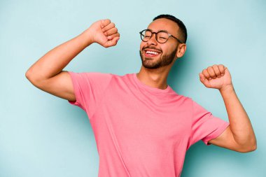 Genç İspanyol adam mavi arka planda izole edilmiş özel bir günü kutluyor, atlıyor ve enerjiyle kollarını kaldırıyor..