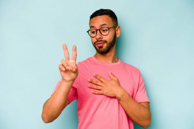 Genç İspanyol bir adam mavi arka planda izole edilmiş yemin ediyor, elini göğsüne koyuyor..