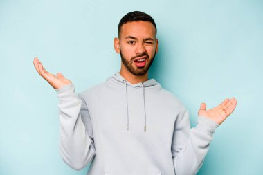 Genç İspanyol bir adam mavi arka planda izole edilmiş. Kafası karışmış ve omuz silkiyor. Kopya alanını tutmak için..