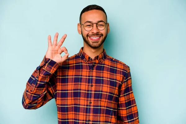 Genç İspanyol adam mavi arka planda izole edilmiş neşeli ve kendine güvenen iyi bir hareket sergiliyor..