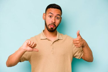 Latin kökenli genç adam mavi arka planda izole edilmiş, baş parmağını kaldırıyor, zor bir seçim konsepti.