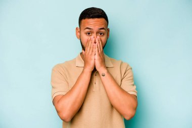 Genç İspanyol bir adam mavi arka planda izole edilmiş, ağzı kapalı ve elleri endişeli görünüyor..