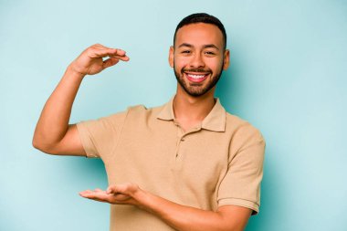Mavi arka planda izole edilmiş genç İspanyol adam işaret parmaklarıyla küçük bir şey tutuyor, gülümsüyor ve kendinden emin..