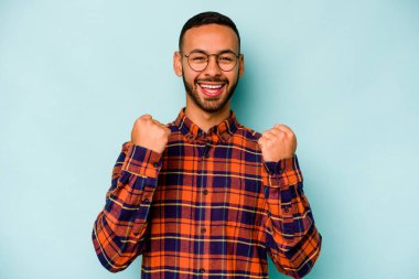 Genç İspanyol adam mavi arka planda izole edilmiş kaygısız ve heyecanlı tezahüratlar yapıyor. Zafer konsepti.