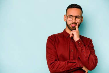 Genç İspanyol adam mavi arka planda izole edilmiş şüpheli ve şüpheci bir ifadeyle yana bakıyor..