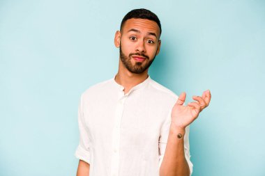 Latin kökenli genç bir adam, mavi arka planda izole edilmiş şüphe içinde ve sorgulama sırasında omuz silkiyor..
