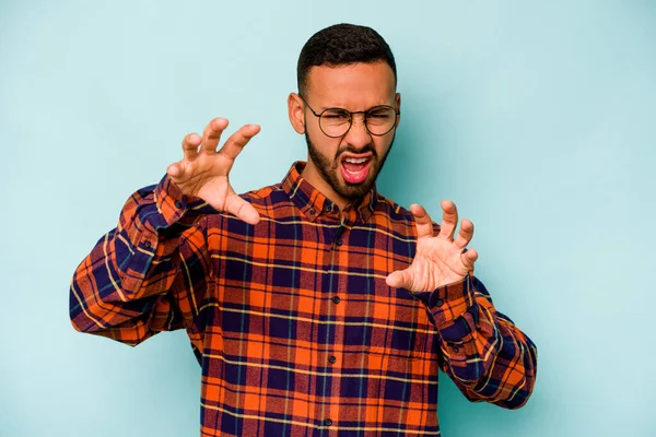 Mavi arka planda izole edilmiş genç İspanyol bir adam pençelerini bir kediyi taklit ederken gösteriyor, agresif bir hareket..