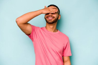 Mavi arka planda izole edilmiş genç İspanyol adam ellerini başının üstünde tutarak neşeyle gülüyor. Mutluluk kavramı.