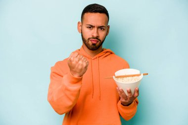 Mavi arka planda izole edilmiş erişte yiyen genç İspanyol kadın kameraya yumruk gösteriyor, agresif yüz ifadesi..