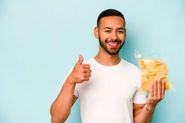 Mladý Hispánec Drží Pytel Chipsů Izolovaných Modrém Pozadí Úsměvem Zvednutím — Stock fotografie