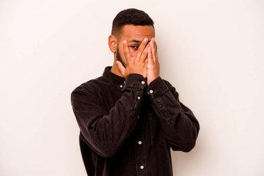 Beyaz arka planda izole edilmiş genç İspanyol adam parmaklarının arasından korkmuş ve gergin bir şekilde göz kırpıyor..