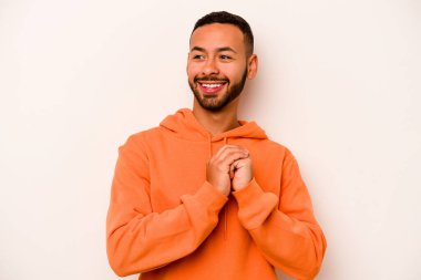 Latin kökenli genç bir adam beyaz arka planda izole edilmiş şans için dua ediyor, şaşırmış ve ağzı açık bir şekilde önüne bakıyor..