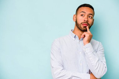 Genç İspanyol adam mavi arka planda izole edilmiş şüpheli ve şüpheci bir ifadeyle yana bakıyor..