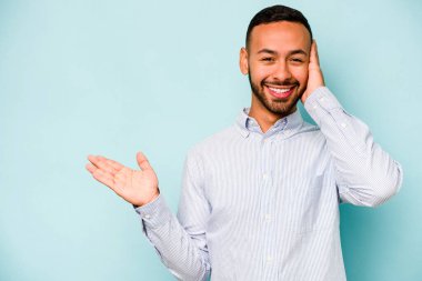 Mavi arka planda izole edilmiş genç İspanyol bir adam avucunun içinde fotokopi alanı tutuyor, el üstünde tutuyor. Şaşırmış ve mutlu..