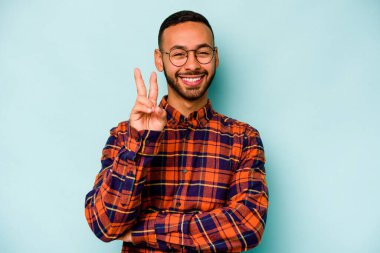 Mavi arka planda izole edilmiş genç İspanyol adam iki numarayı parmaklarıyla gösteriyor..