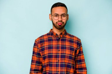 Mavi arka planda izole edilmiş genç İspanyol adam yanakları patlatıyor, yorgun bir ifadesi var. Yüz ifadesi kavramı.