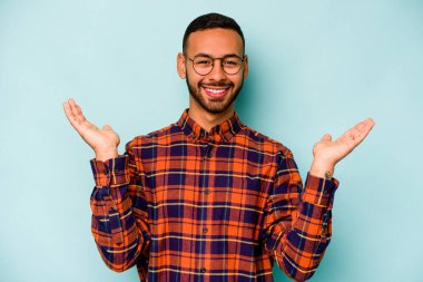 Mavi arka planda izole edilmiş genç İspanyol adam kollarıyla ölçeği yapıyor, mutlu ve kendinden emin hissediyor..