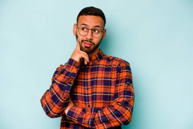 Latin kökenli genç bir adam mavi arka planda izole edilmiş. Bir strateji planlıyor, bir iş yolu düşünüyor..