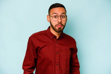 Mavi arka planda izole edilmiş genç bir İspanyol omuz silker ve gözleri açık kalır..