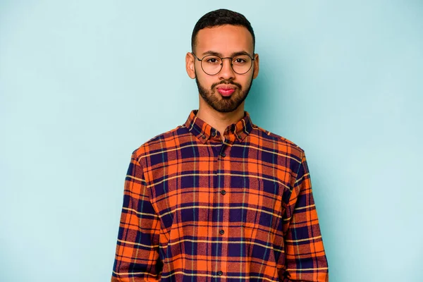 Mavi arka planda izole edilmiş genç İspanyol adam yanakları patlatıyor, yorgun bir ifadesi var. Yüz ifadesi kavramı.