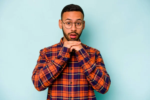 Latin kökenli genç adam mavi arka planda izole edilmiş şans için dua ediyor, şaşırmış ve ağzı açık bir şekilde önüne bakıyor..