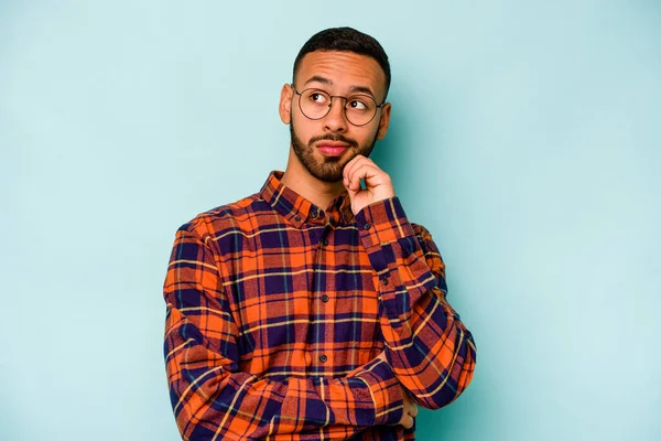 Genç İspanyol adam mavi arka planda izole edilmiş düşünüyor, bakıyor, yansıtıyor, düşünüyor, hayal kuruyor..