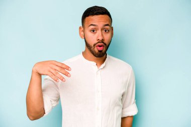 Genç İspanyol bir adam mavi arka planda izole edilmiş bir şekilde bir şeye gülüyor, elleriyle ağzını kapatıyor..