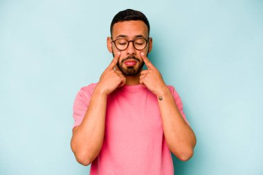 Genç İspanyol bir adam mavi arka planda izole edilmiş, ağlıyor, bir şeyden memnun değil, acı ve kafa karışıklığı konsepti..