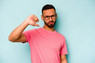 Mavi arka planda izole edilmiş genç İspanyol adam baş parmağını aşağı gösteriyor ve hoşlanmadığını ifade ediyor..