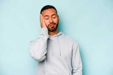 Genç İspanyol adam mavi arka planda izole edilmiş. Çok yorgun ve uykulu. Elini başının üstünde tutuyor..