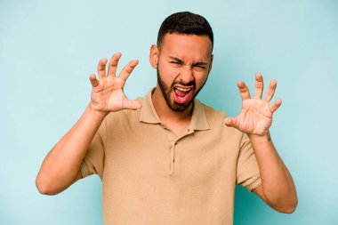 Mavi arka planda izole edilmiş genç İspanyol bir adam pençelerini bir kediyi taklit ederken gösteriyor, agresif bir hareket..