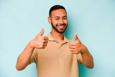 Genç İspanyol adam mavi arka planda izole edilmiş. Baş parmağını kaldırıyor, gülümsüyor ve kendinden emin..