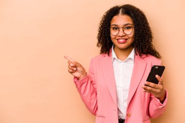 Elinde cep telefonu olan genç Afrikalı bir kadın bej arka planda izole edilmiş gülümseyen ve işaret eden boş alanda bir şey gösteriyor..