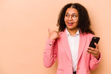 Elinde cep telefonu tutan genç Afrikalı bir kadın. Bej arka plan noktalarında izole edilmiş. Baş parmak uzakta, gülen ve kaygısız..